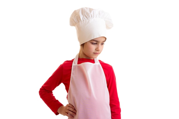 Niña encantadora con camisa roja con delantal blanco y sombrero de cocina mirando hacia abajo en el estudio