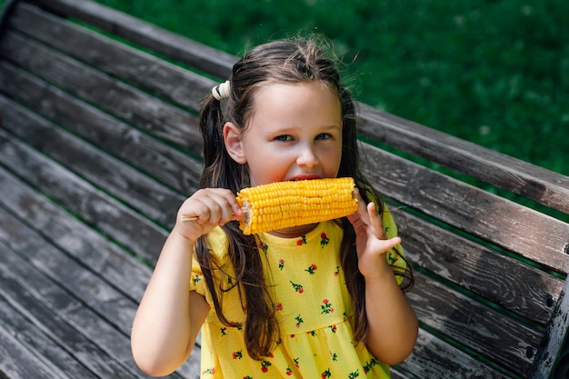 Una niña encantadora come deliciosas vacaciones familiares de maíz dulce hervido el fin de semana en el parque de la ciudad