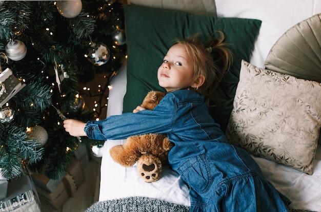 Una niña encantadora se encuentra con su osito de peluche bajo el árbol de Navidad