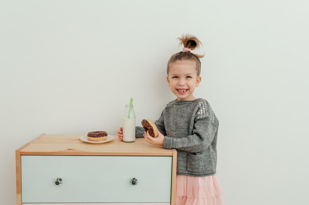 Una niña encantadora está comiendo donas de chocolate con una botella de leche