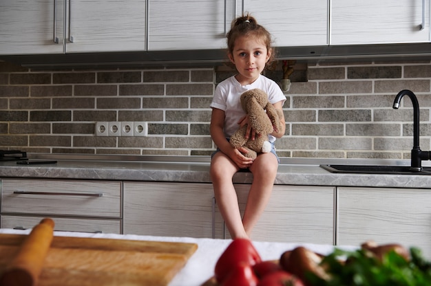 Una niña encantadora está sentada en la encimera de la cocina con un peluche en sus manos. Verduras en la mesa en primer plano