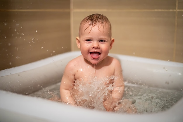 Niña encantadora juega en el agua en una pequeña piscina inflable para bebés ubicada en la ducha en el baño, niña sonriendo y divirtiéndose