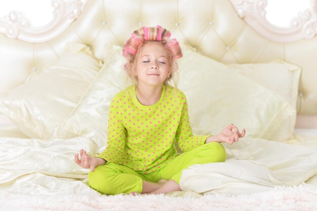 Niña encantadora con rulos rosas meditando en la cama en casa