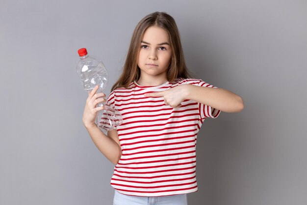 Una niña encantadora y seria con camiseta a rayas sosteniendo y señalando con el dedo una botella de plástico vacía