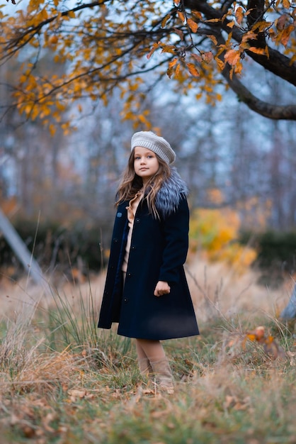 Una niña encantadora con un sombrero y un abrigo azul está parada en un jardín de otoño