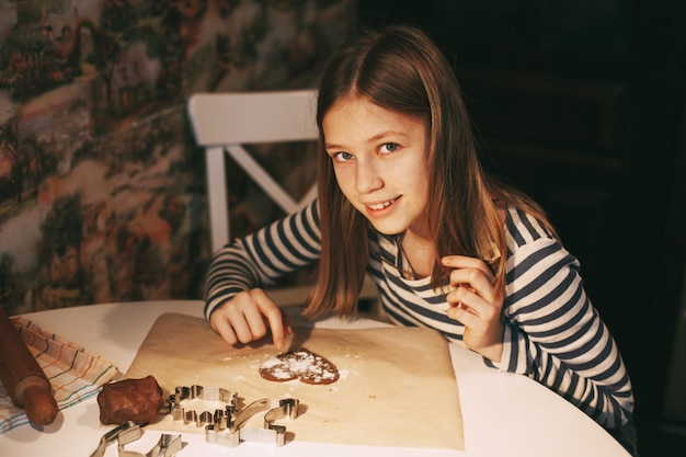 Una niña encantadora y sonriente en la cocina de la casa, en la mesa, cortó galletas con forma de corazón de la masa