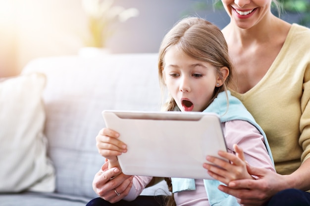 Niña encantadora y su hermosa madre joven con tableta digital y sonriendo mientras está sentado en el sofá en casa