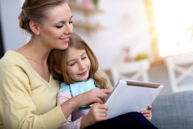 Niña encantadora y su hermosa madre joven con tableta digital y sonriendo mientras está sentado en el sofá en casa