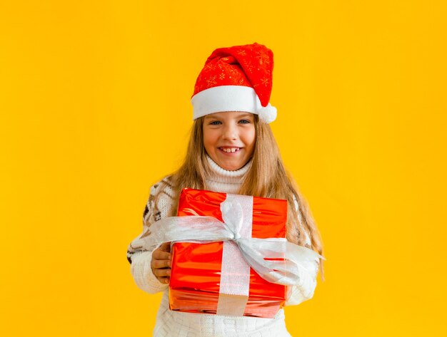 Niña encantadora con un suéter de punto y gorro de Papá Noel con un regalo sobre un fondo amarillo.