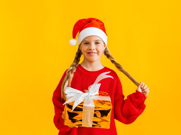 Niña encantadora con un suéter de punto y gorro de Papá Noel con un regalo sobre un fondo amarillo.