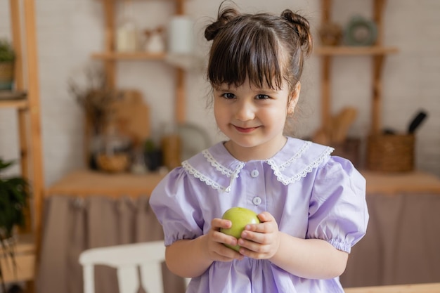 una niña encantadora con un vestido lila come una manzana verde en la cocina. espacio para texto, pancarta