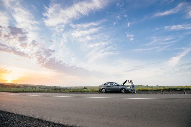 Una niña se encuentra cerca de un automóvil averiado en medio de la carretera durante el atardecer e intenta pedir ayuda por teléfono. Avería y reparación del coche. Esperando ayuda.