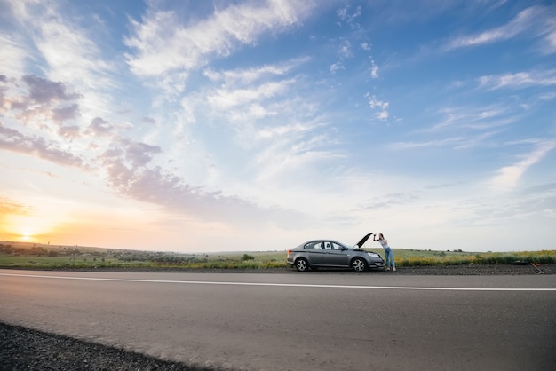 Una niña se encuentra cerca de un automóvil averiado en medio de la carretera durante el atardecer e intenta pedir ayuda por teléfono. Avería y reparación del coche. Esperando ayuda.