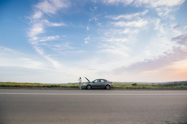 Una niña se encuentra cerca de un automóvil averiado en medio de la carretera durante el atardecer e intenta pedir ayuda por teléfono. Avería y reparación del coche. Esperando ayuda.