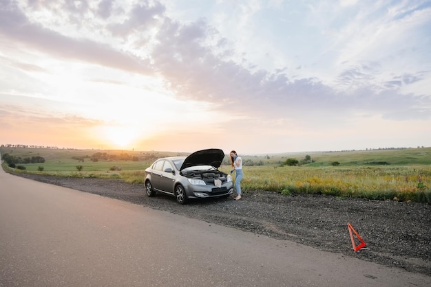 Una niña se encuentra cerca de un automóvil averiado en medio de la carretera durante el atardecer e intenta repararlo. Avería y reparación del coche. Solución de problemas del problema.