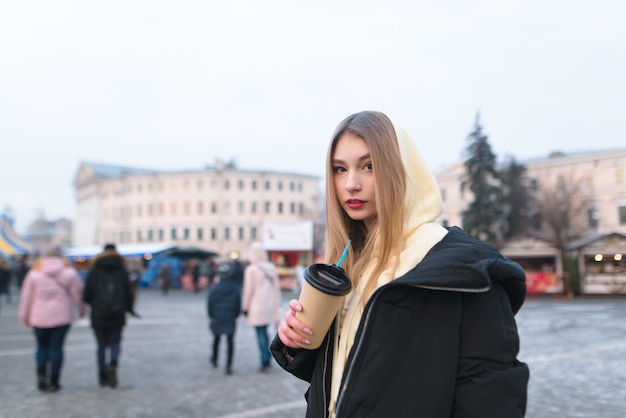 Niña se encuentra en el fondo de la calle de invierno de una ciudad con una taza de café