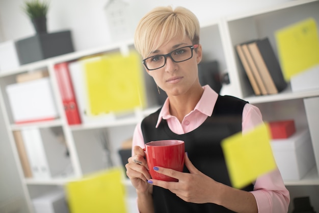 Una niña se encuentra en la oficina cerca de un tablero transparente con calcomanías y sostiene una taza roja en la mano.