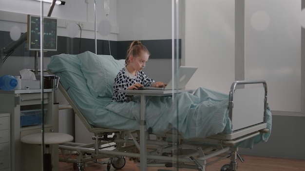 Niña enferma hospitalizada con tubo nasal de oxígeno sentada en la cama jugando videojuegos en línea en la computadora portátil mientras se recupera después de una cirugía médica. Niño esperando tratamiento durante la consulta por enfermedad