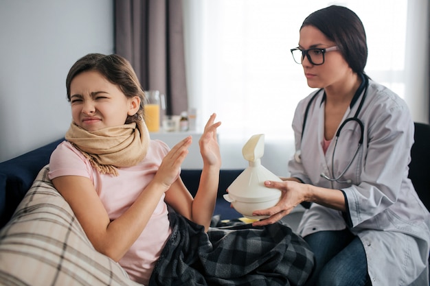 La niña enferma obstinada se encoge y mantiene los ojos cerrados. Ella no quiere hacer un procedimiento de inhalación. Doctora sostenga el inhalador blanco y déselo al niño. Se sientan en una habitación.