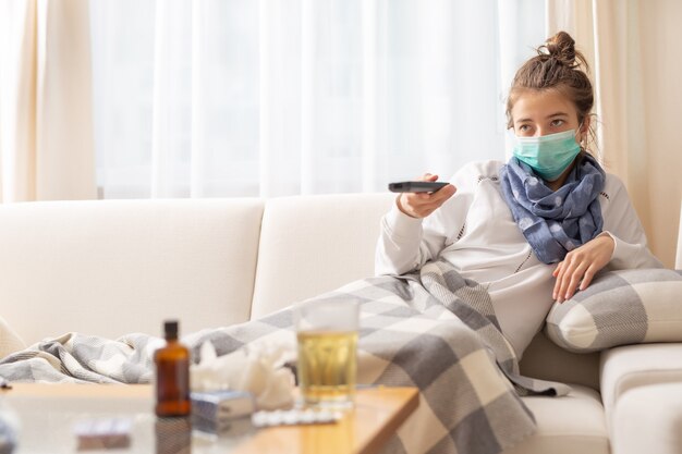 Niña enferma sentada en un sofá en casa viendo la televisión. Tiene neumonía por coronavirus y usa una máscara protectora respiratoria de seguridad para protegerse a sí misma y a los demás de las enfermedades.