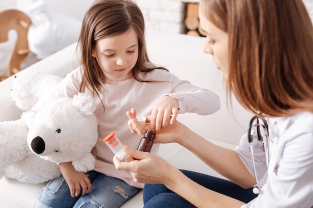 Niña enferma tomando pastillas mientras un médico profesional la ayuda mientras está sentada en el sofá juntos