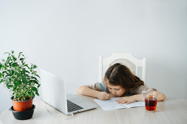 Niña enfocada está estudiando escribir en un cuaderno frente a una computadora portátil