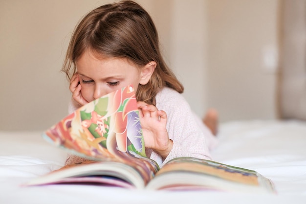 Niña enfocada leyendo un libro en la cama Día internacional de la alfabetización