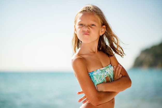 Niña enojada parada en la playa con los brazos cruzados y frunciendo el ceño.