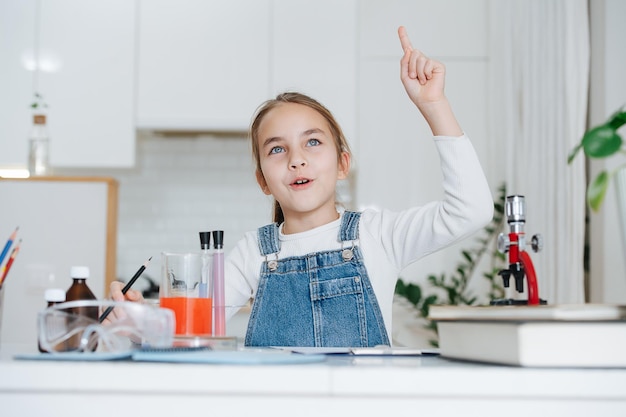 Niña entusiasta haciendo proyecto de ciencia en casa, teniendo un momento eureka. Ella tiene cristalería de química con líquidos coloridos y microscopio.