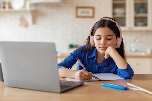 Niña escolar aburrida aprendiendo en la computadora portátil tomando notas en casa