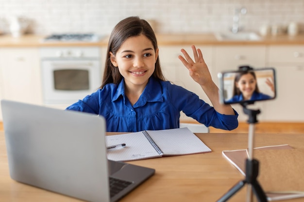 Foto niña escolar sentada cerca de la computadora portátil saludando al teléfono interior