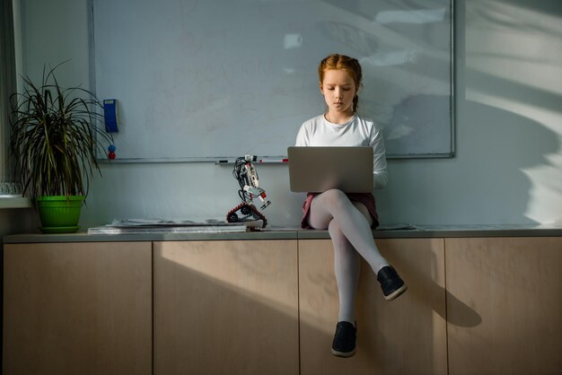 Niña escolar usando una computadora portátil en la mesa en el aula con un robot DIY