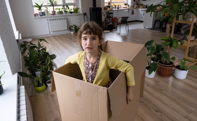 La niña se esconde en una caja grande y se ve divertida en una habitación dentro de la casa Mudarse a una casa nueva desempacar cosas alquilar un apartamento hipoteca problema de vivienda