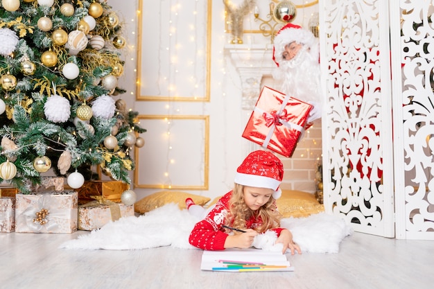 La niña escribe una carta y Santa Claus con un regalo la espía en el árbol de Navidad