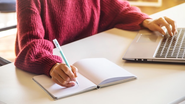 La niña escribe en un cuaderno y trabaja en su computadora portátil sobre una mesa blanca en brillante coworking.