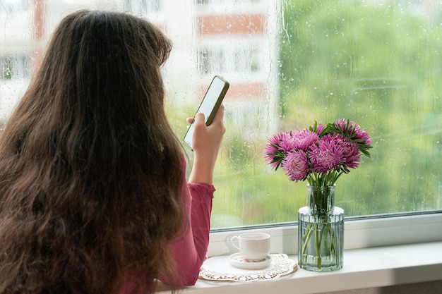 la niña escribe sms en un teléfono móvil junto a una ventana lluviosa.