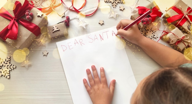 Foto niña escribiendo una carta de navidad a santa en papel blanco sobre fondo de madera con adornos navideños.