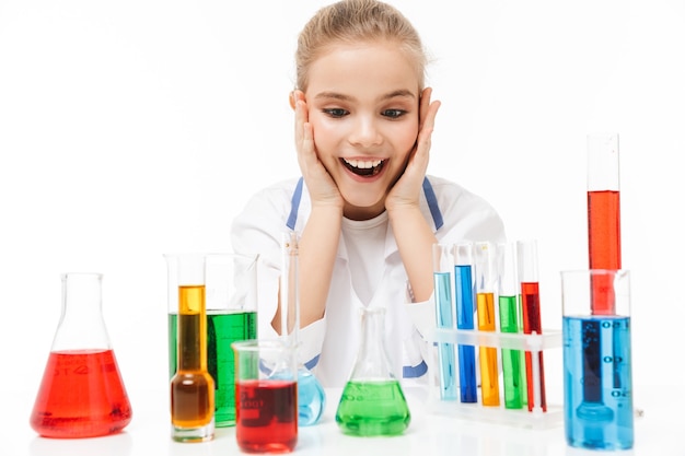 Niña de la escuela alegre en bata blanca de laboratorio haciendo experimentos químicos con líquido multicolor en tubos de ensayo aislado sobre pared blanca