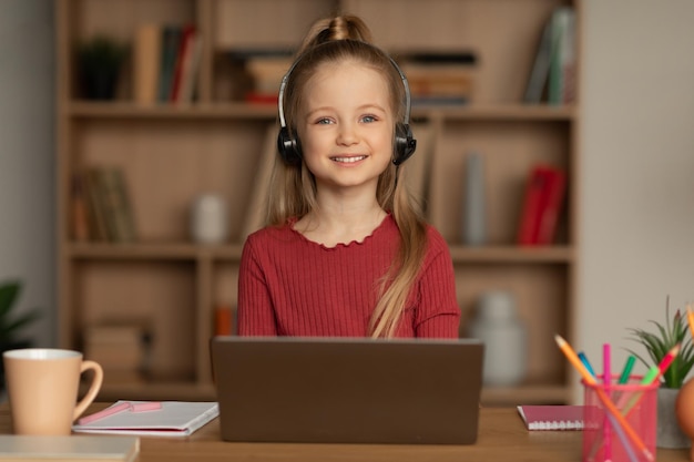 Niña de la escuela alegre que usa una computadora portátil con auriculares en casa