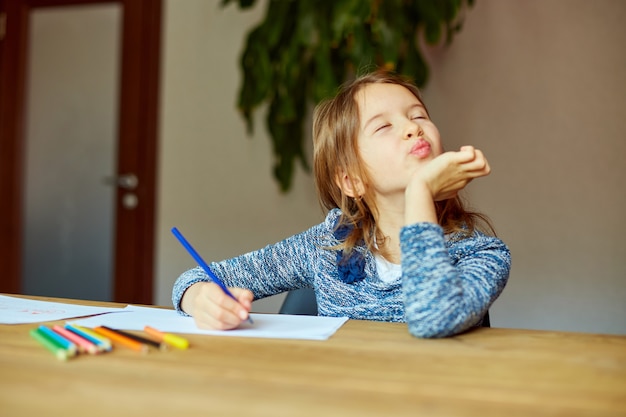 Niña de la escuela dibujando y escribiendo una imagen con crayones, usando lápices de colores en la mesa en casa