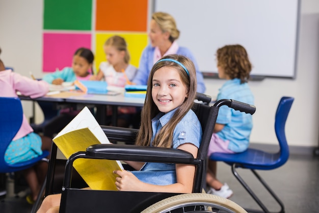 Niña de la escuela con discapacidad en silla de ruedas sosteniendo un libro en el aula