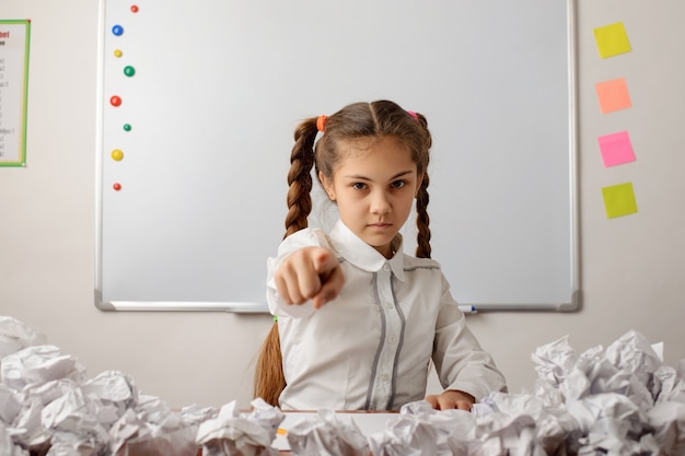 Niña de la escuela enojada apuntando a la cámara, sentada en el escritorio con muchos papeles arrugados alrededor