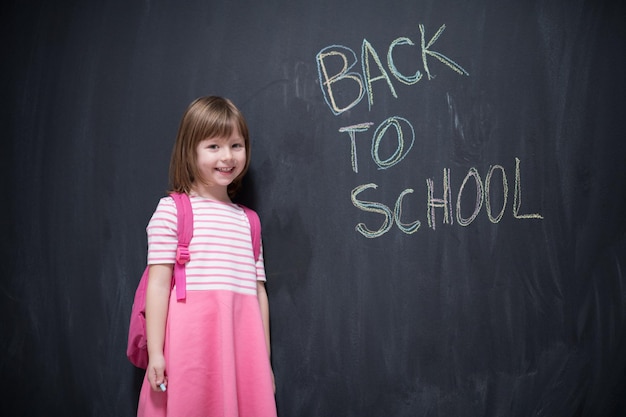 Niña de la escuela feliz con mochila escribiendo de regreso a la escuela en pizarra negra