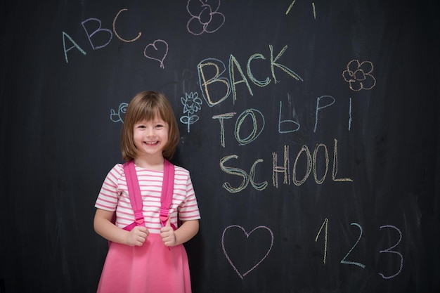 Niña de la escuela feliz con mochila escribiendo de regreso a la escuela en pizarra negra