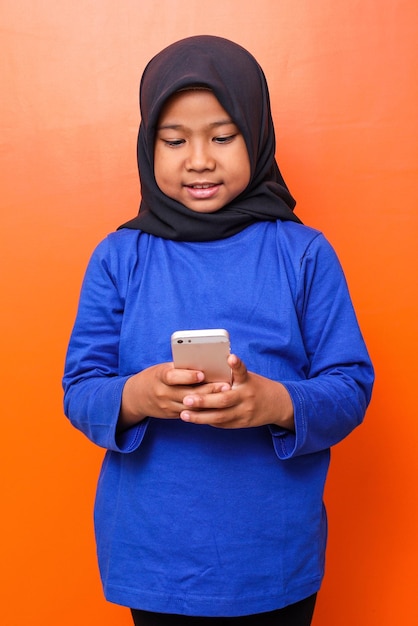 Niña de la escuela musulmana sonriendo mientras juega al teléfono inteligente contra el fondo naranja