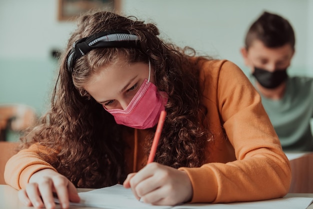 Foto niña de la escuela de niños caucásicos con mascarilla estudiando en el aula