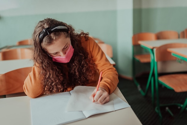 Niña de la escuela de niños caucásicos con mascarilla estudiando en el aula