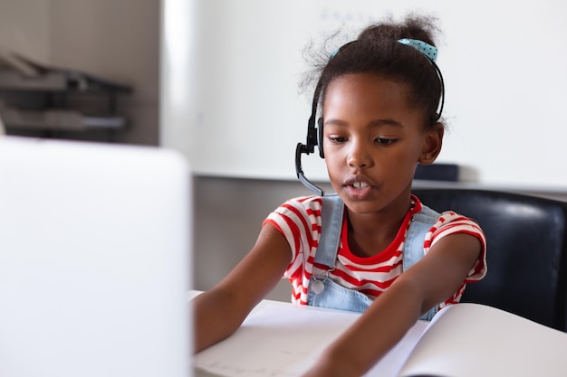 Niña de escuela primaria afroamericana con auriculares mientras usa una computadora portátil en el escritorio en clase