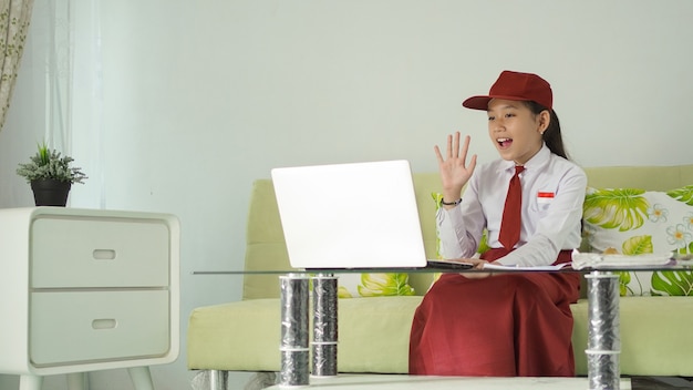 Niña de la escuela primaria asiática que estudia desde casa saludando a la pantalla de su computadora portátil