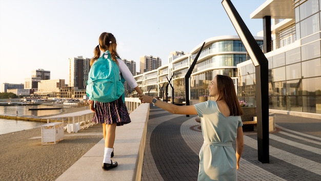 Niña de la escuela en uniforme está caminando sobre la barandilla del paseo marítimo mientras sostiene la mano de su joven madre